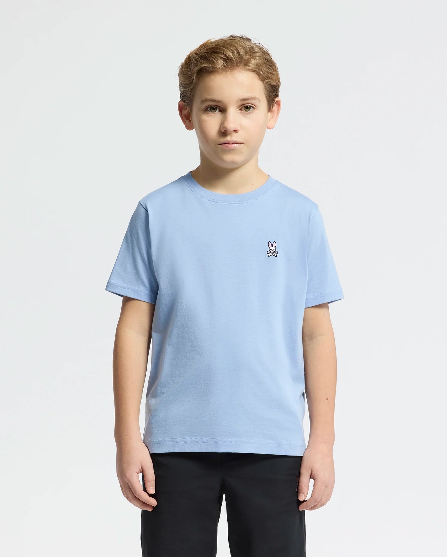 A young boy with short hair stands against a plain background wearing the Psycho Bunny KIDS CLASSIC CREW NECK TEE in light blue, crafted from soft Pima cotton with an embroidered Bunny logo on the chest, paired with sleek black pants.
