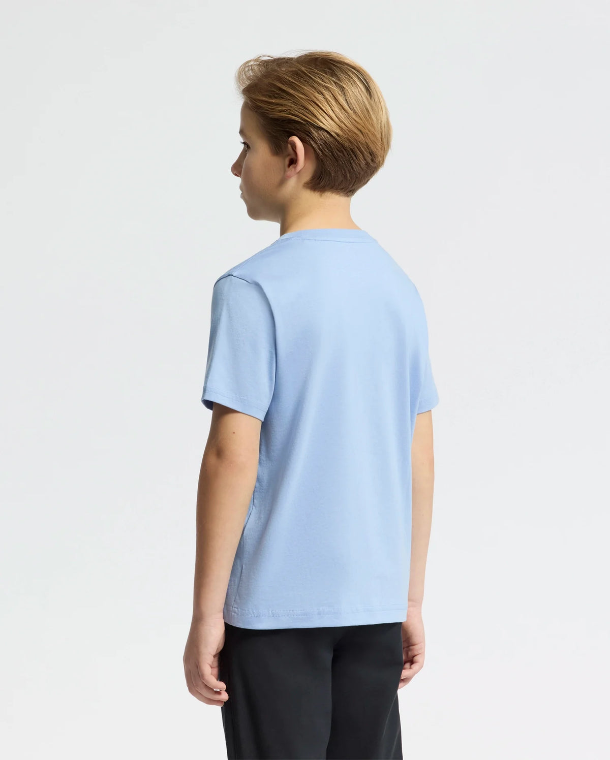 A young boy with short, light brown hair stands facing away, wearing the KIDS CLASSIC CREW NECK TEE - B0U014B200 by Psycho Bunny in light blue, featuring an embroidered Bunny logo. He pairs it with dark pants against a plain white background.