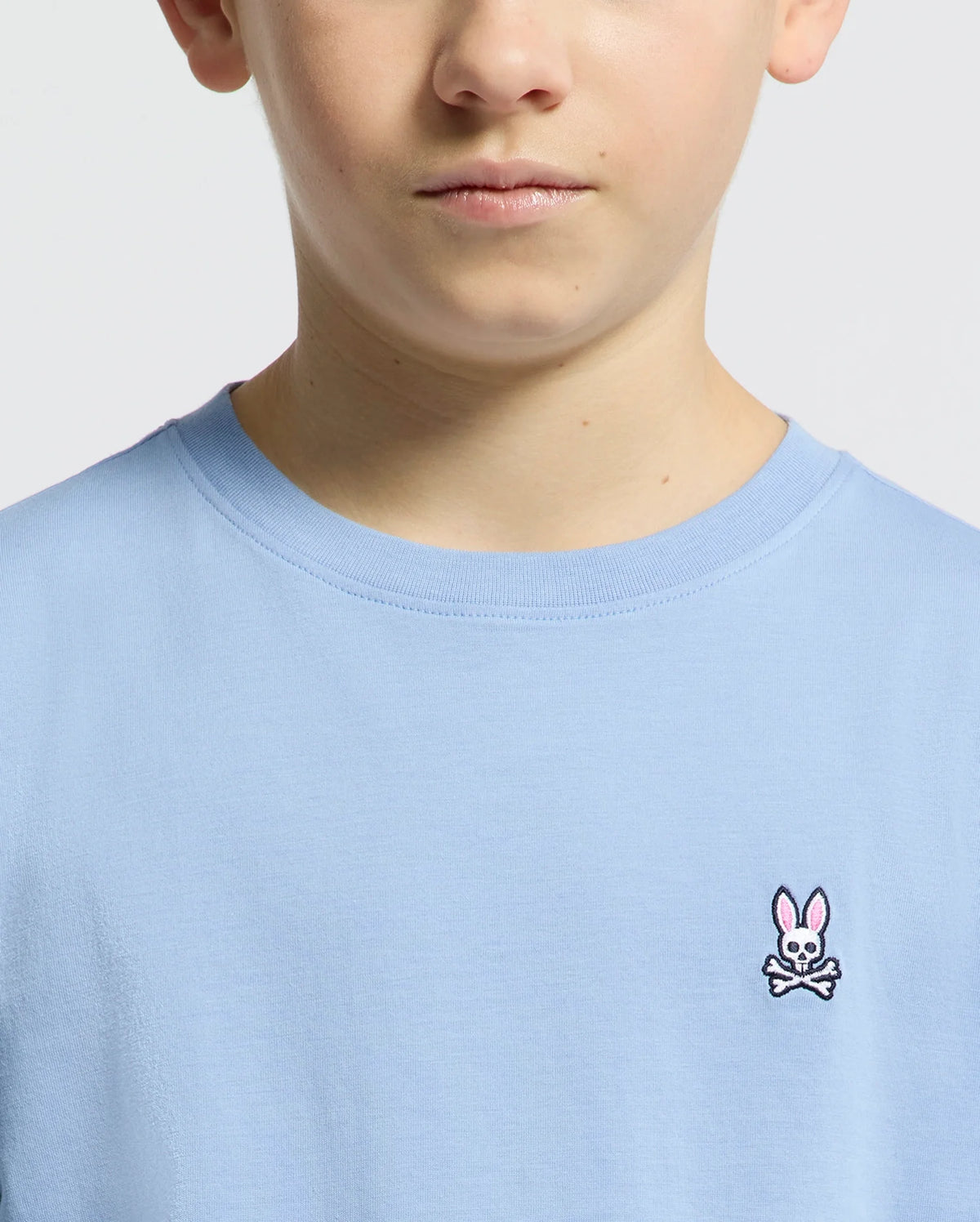 A child wears a Psycho Bunny light blue KIDS CLASSIC CREW NECK TEE (B0U014B200) made of Pima cotton, featuring an embroidered bunny logo with crossed bones. The focus is on the neck and upper chest area.