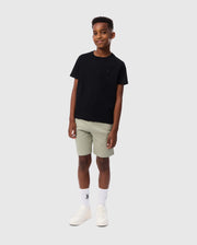 A young boy stands against a plain background, wearing the Psycho Bunny Kids Embroidered Classic Tee (B0U020AR00) made from luxurious Peruvian Pima cotton. He pairs it with light gray shorts, white socks, and white sneakers. His relaxed and confident pose, along with his slight smile, completes the timeless silhouette.