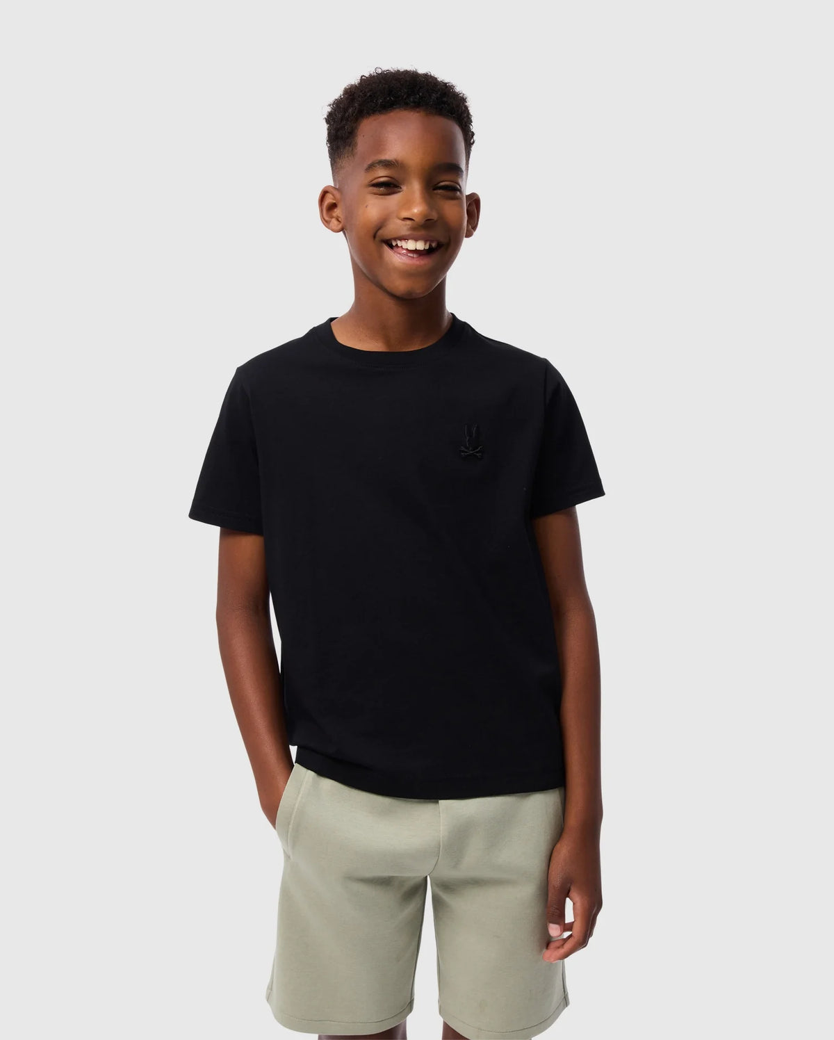 A young boy is smiling and standing against a plain background. He is wearing a Psycho Bunny KIDS EMBROIDERED CLASSIC TEE - B0U020AR00 in black and light gray shorts, with his hands in his pockets.