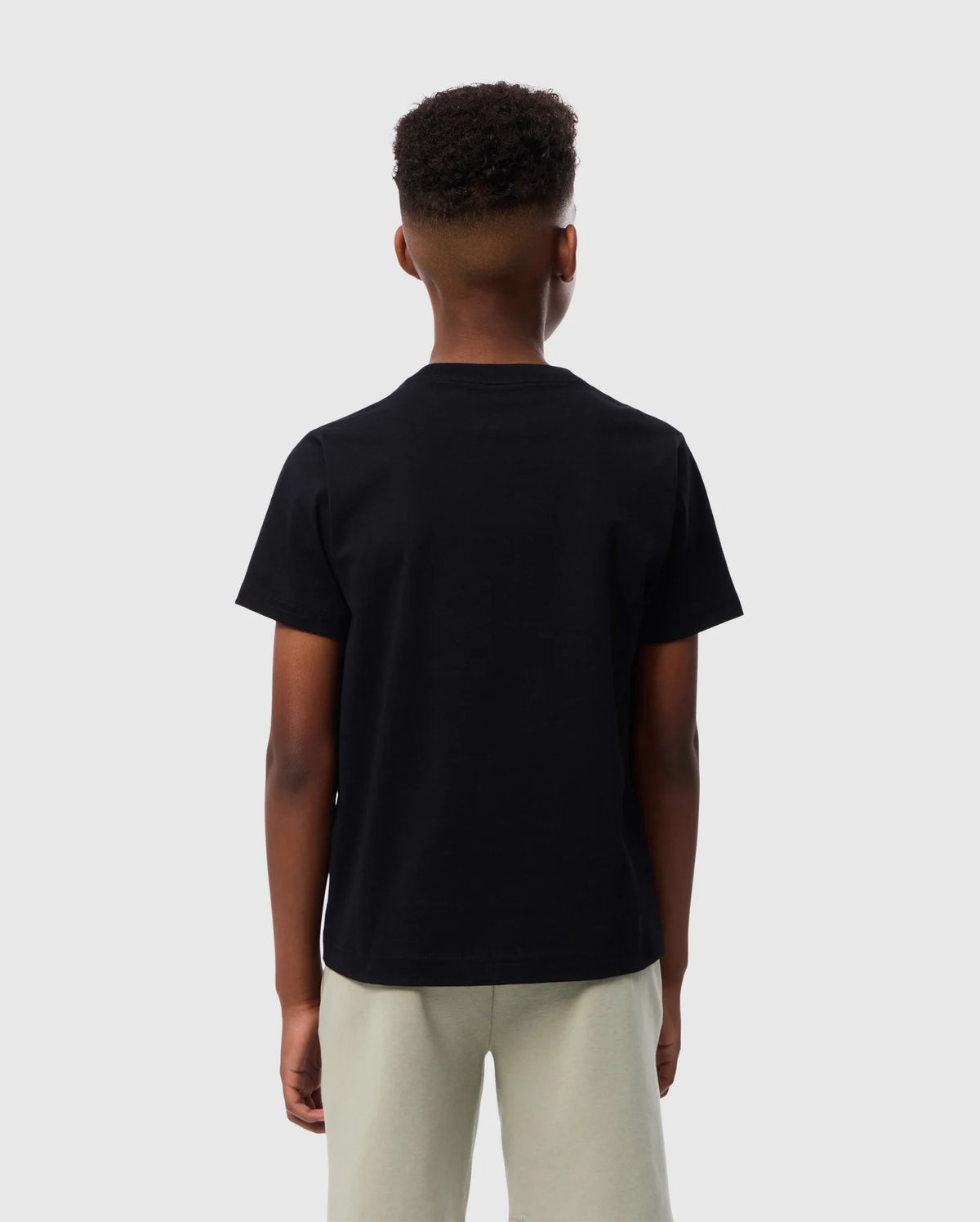 A person wearing a plain black KIDS EMBROIDERED CLASSIC TEE - B0U020AR00 from Psycho Bunny and light-colored pants is standing with their back to the camera. The background is a solid, light grey color. The person's hair is short and closely clipped on the sides, with more volume on top.