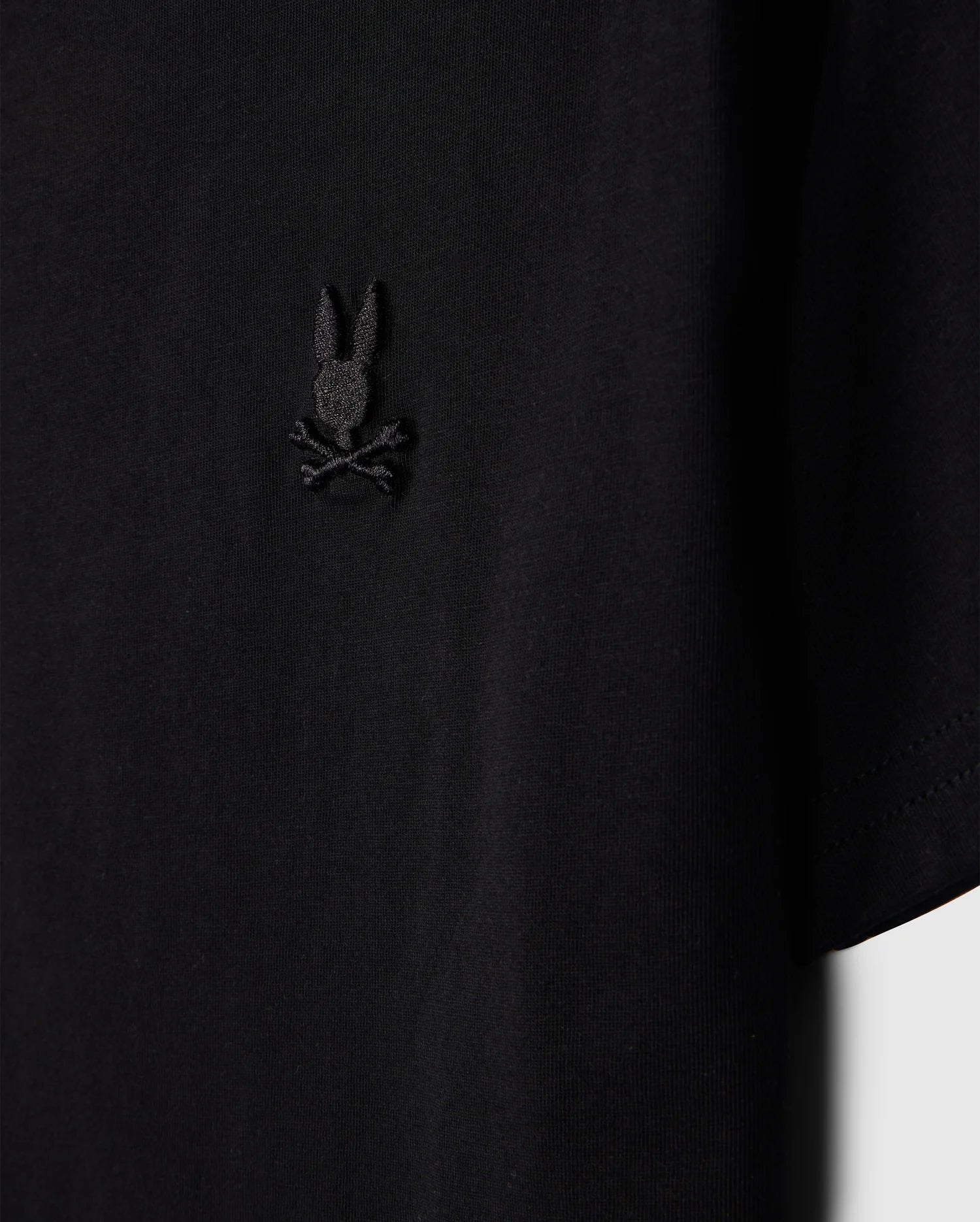 Close-up of a Psycho Bunny Kids Embroidered Classic Tee, product code B0U020AR00, crafted from luxurious Peruvian Pima cotton. It features a small bunny head with crossed bones design on the left chest and is displayed against a plain background for a timeless silhouette.