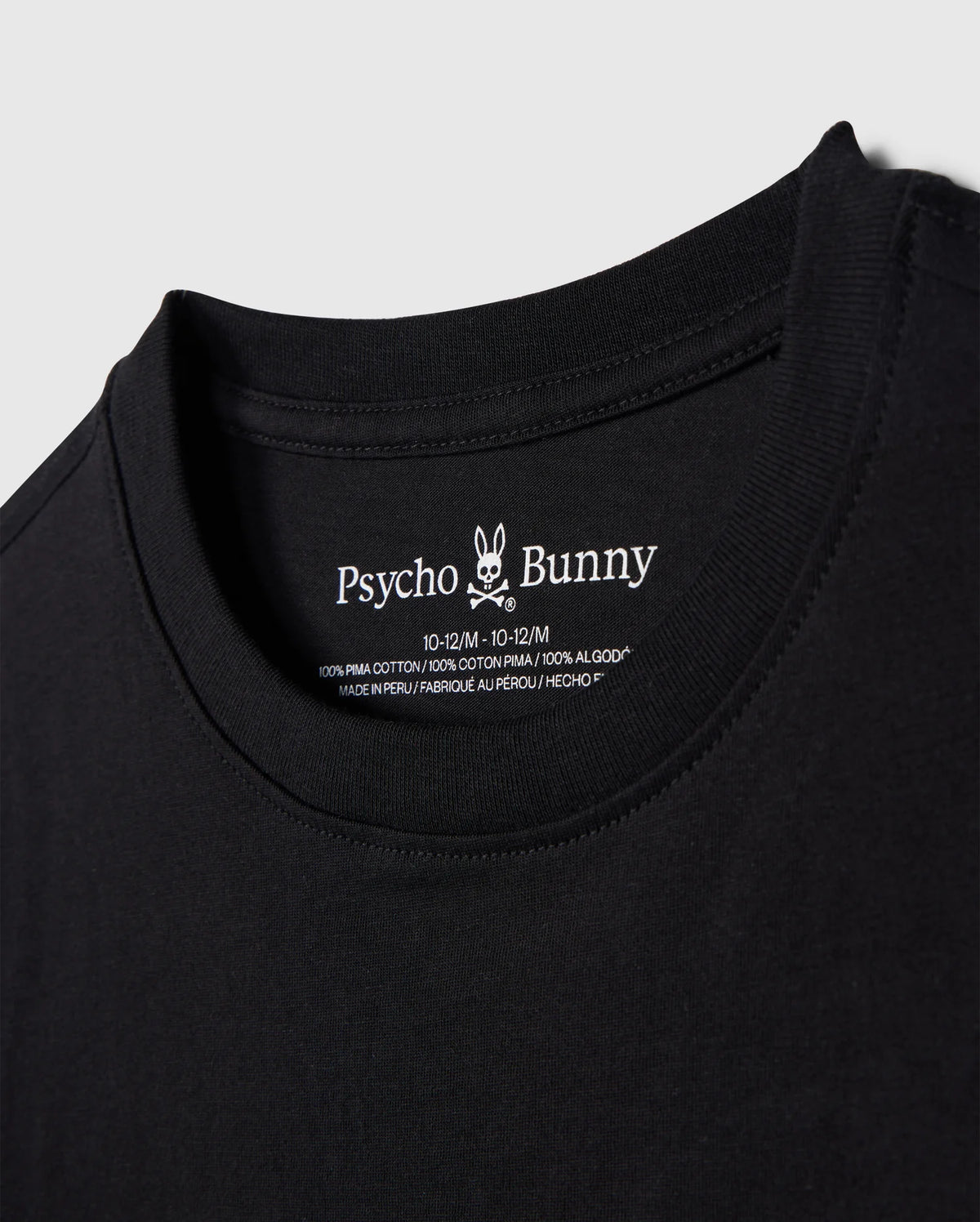 Close-up image of a black T-shirt neckline with a label showing the brand name "Psycho Bunny" alongside its bunny logo, and the size 10-12 M. The KIDS EMBROIDERED CLASSIC TEE - B0U020AR00 is made of 100% Peruvian Pima cotton, ensuring ultimate comfort and quality. This piece showcases a timeless silhouette and is proudly made in Peru.