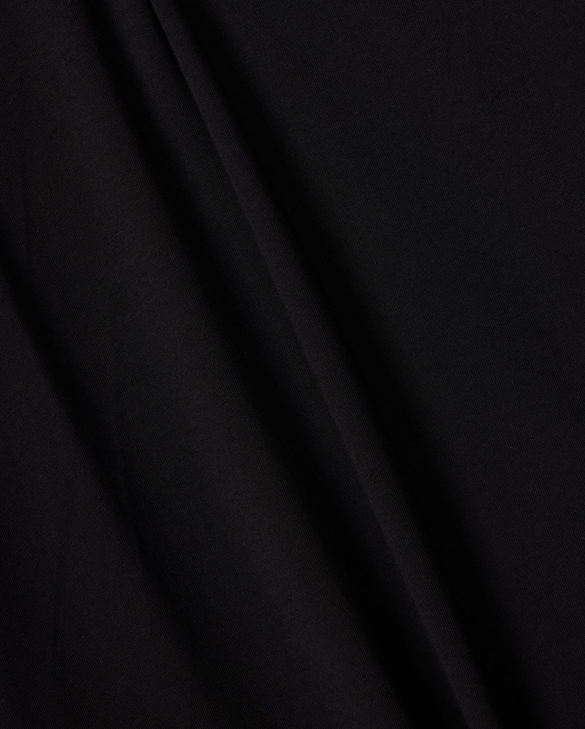 A close-up image of the smooth, black Peruvian Pima cotton fabric used in the KIDS EMBROIDERED CLASSIC TEE - B0U020AR00 by Psycho Bunny. The texture appears soft and slightly matte, with subtle folds and creases creating faint shadows that emphasize its draping and natural curvature, evoking a timeless silhouette.