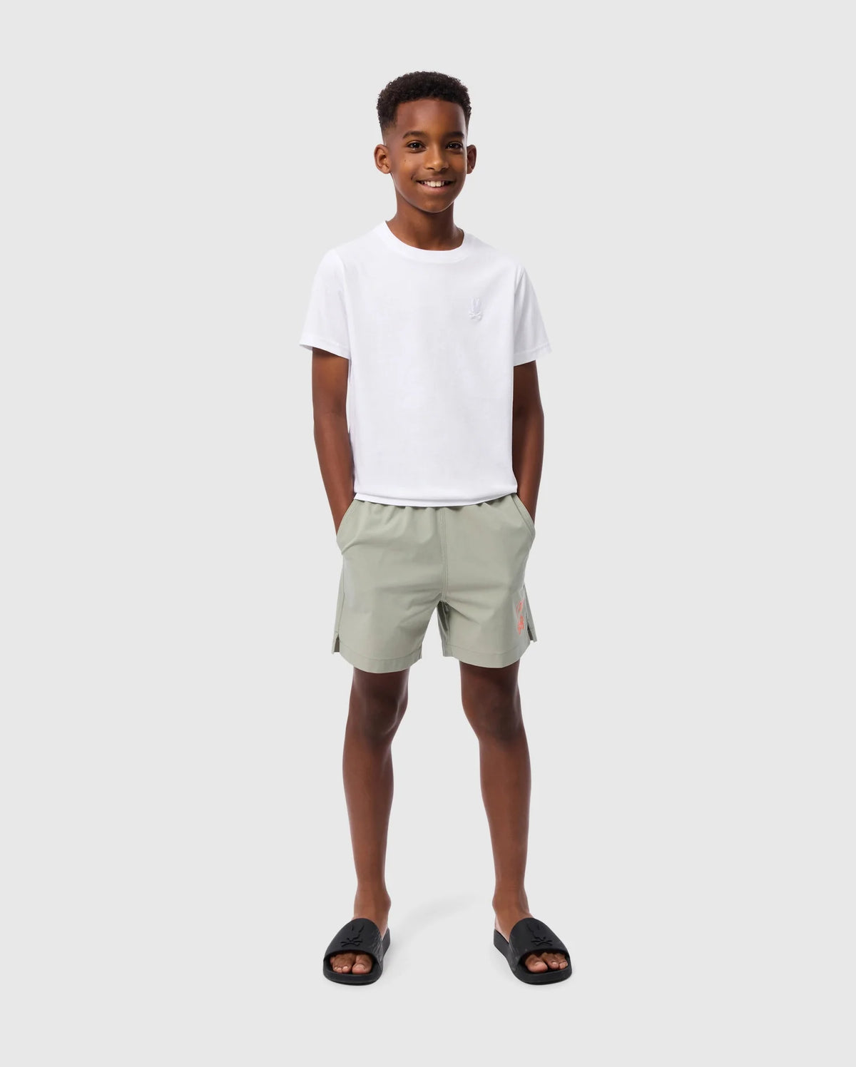 A young boy stands smiling against a plain light grey background. He is wearing the Psycho Bunny KIDS EMBROIDERED CLASSIC TEE - B0U020AR00, made from Peruvian Pima cotton, along with light green shorts and his hands in pockets. His tee features an embroidered chest bunny, adding a playful touch to his relaxed and happy demeanor. Black slides complete his look.