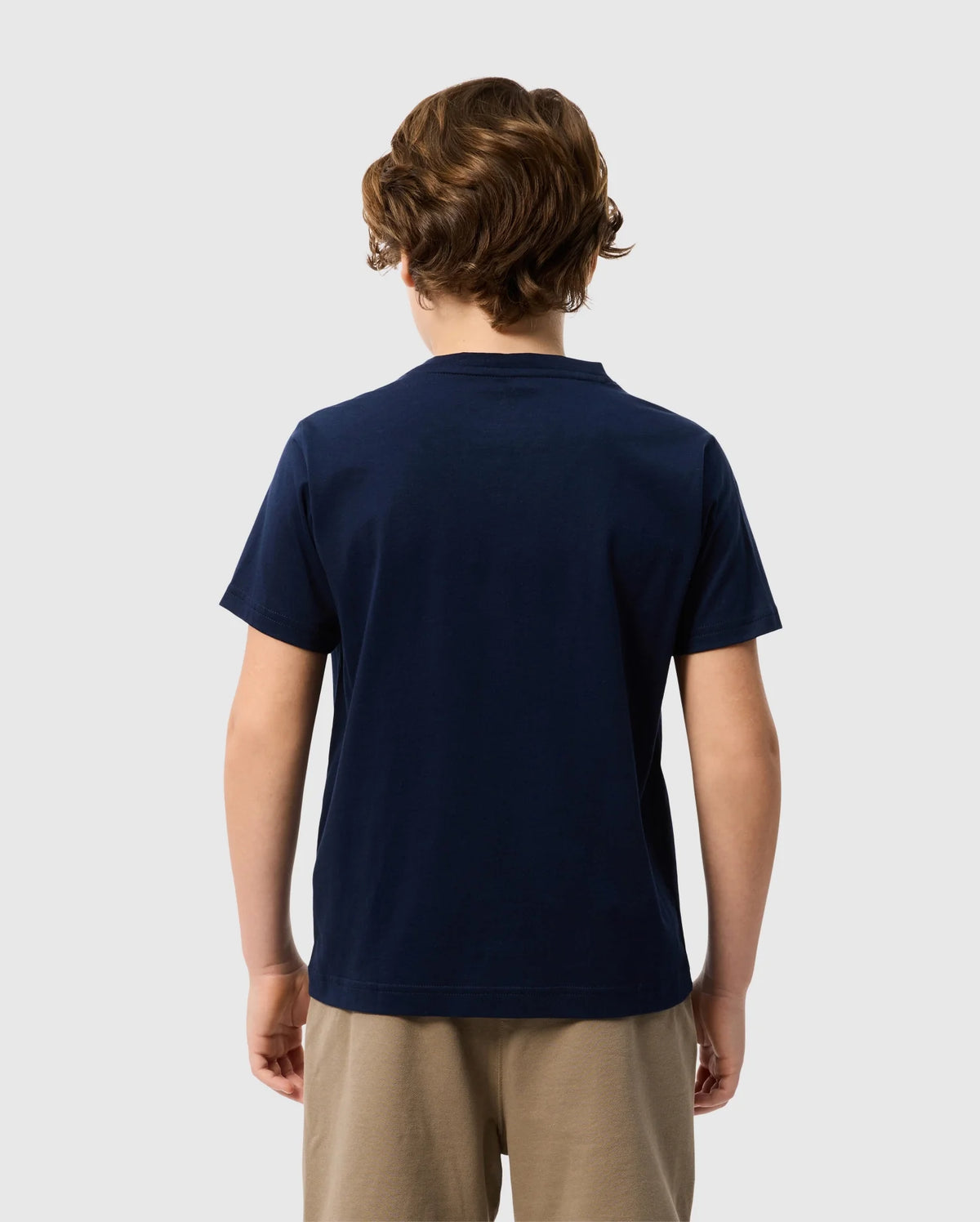 A person with wavy brown hair stands facing away, wearing a navy blue Kids Classic V Neck Tee by Psycho Bunny, made from soft Pima cotton, and beige pants against a plain white background.