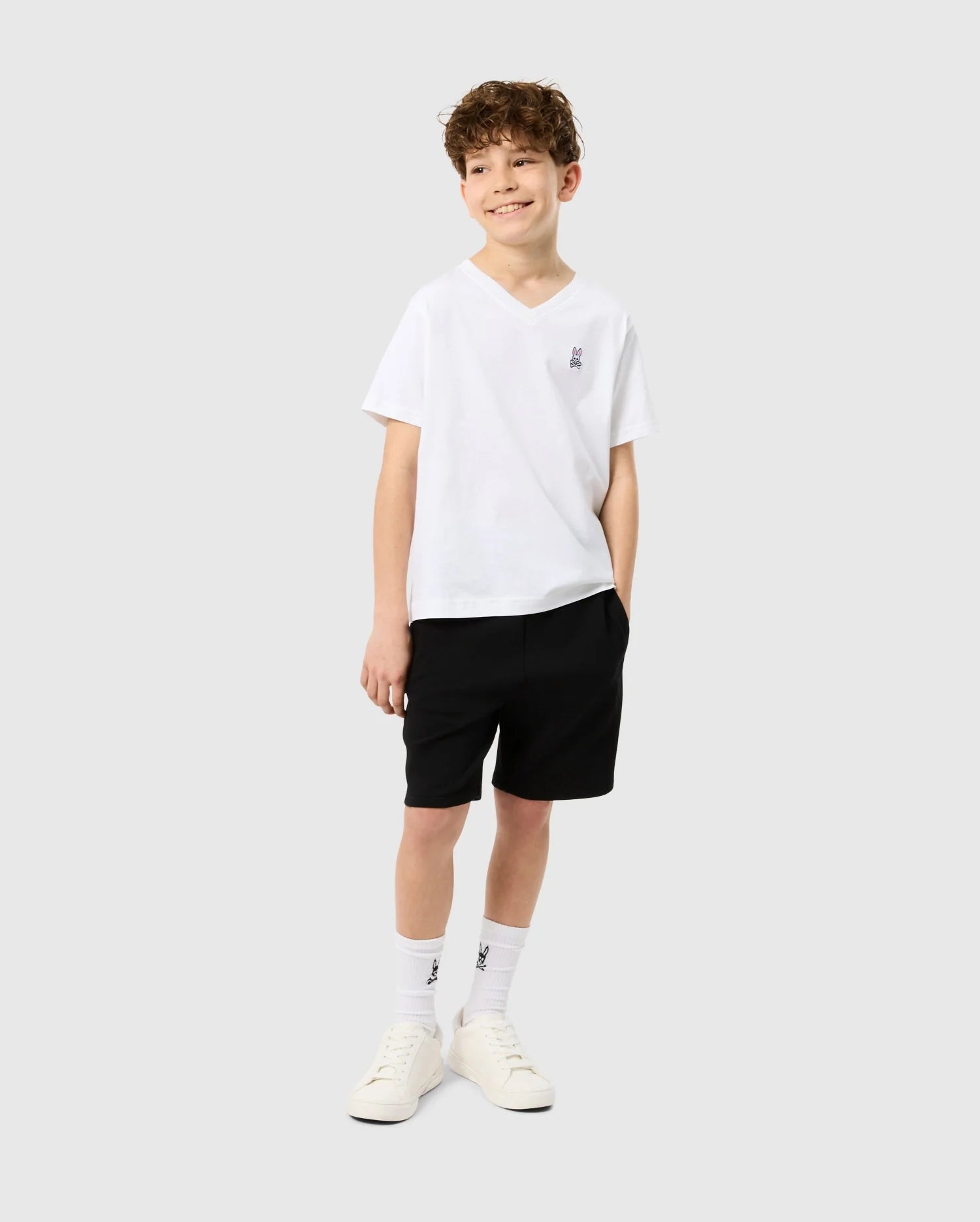 A young boy strikes a pose against a light gray backdrop in a chic white KIDS CLASSIC V NECK TEE - B0U100CRPC by Psycho Bunny, paired with black shorts and white sneakers. Sporting logo-embellished white socks and hands tucked into his pockets, he exudes happiness and readiness for the back-to-school season.