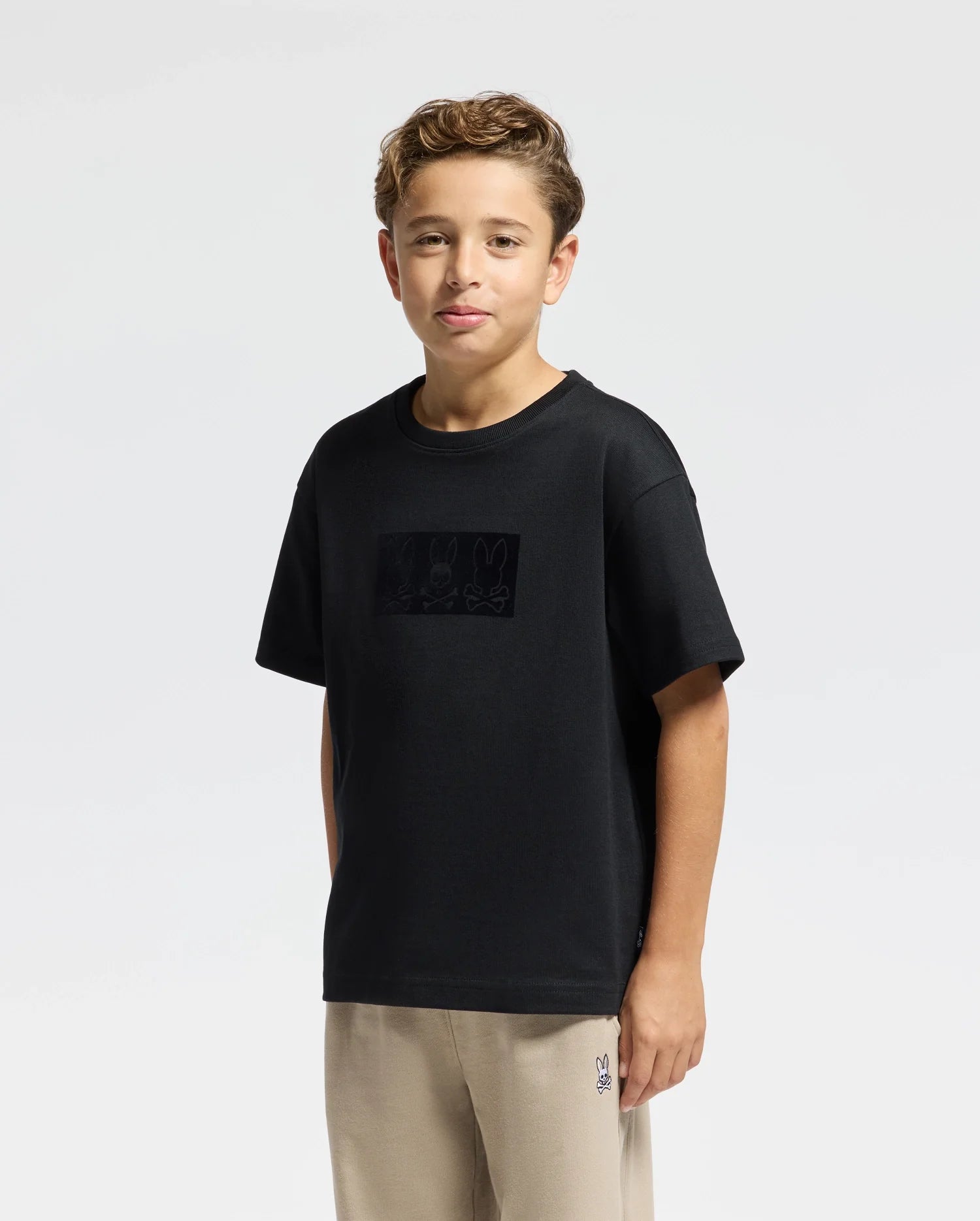 A young boy with curly hair stands against a plain white background wearing a Kids' Romeo Heavy Weight Relaxed Fit Tee by Psycho Bunny, made from soft Pima Cotton. Beige pants complete his look as he rests his hands at his sides, maintaining a neutral expression.