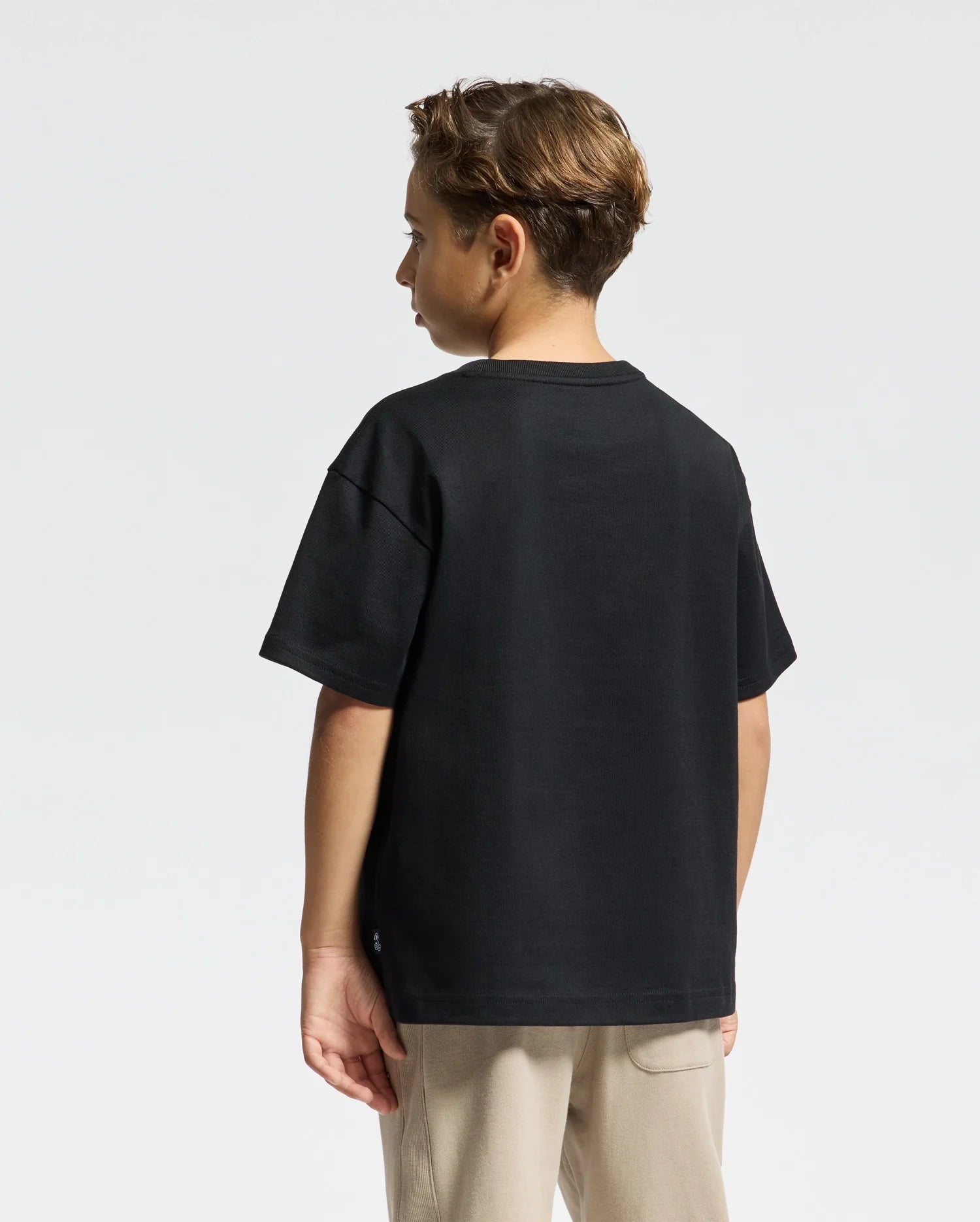 A young individual with short hair stands to the side in a stylish Psycho Bunny KIDS ROMEO HEAVY WEIGHT RELAXED FIT TEE in black and beige pants, exuding casual elegance. The Pima cotton fabric adds a touch of luxury against a plain, light-colored background.