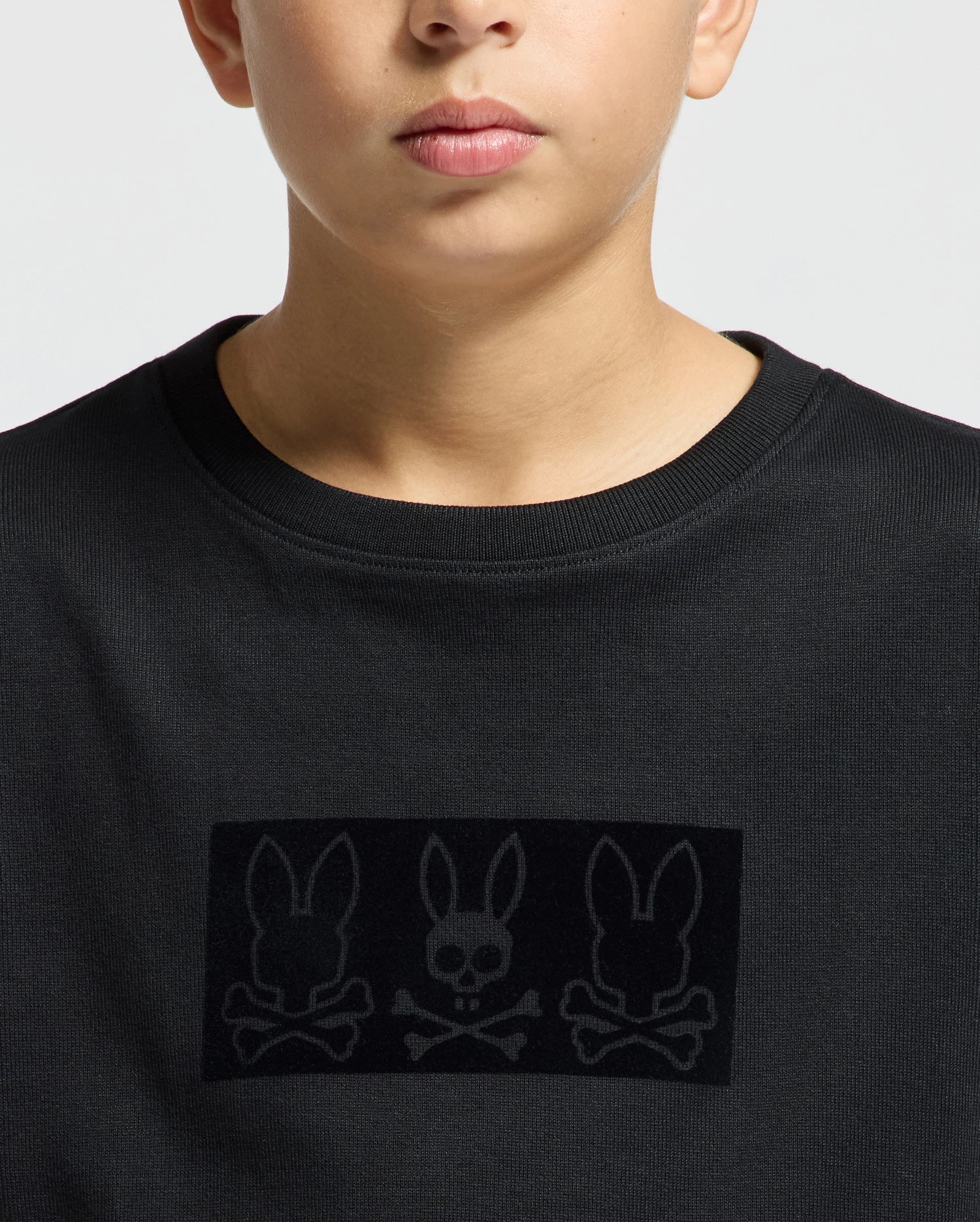 A person wearing the Psycho Bunny KIDS ROMEO HEAVY WEIGHT RELAXED FIT TEE - B0U110E200, which is a black heavyweight tee showcasing a striking design of three rabbit skulls and crossbones arranged in a rectangular pattern across the chest. The simple backdrop enhances the bold graphic.