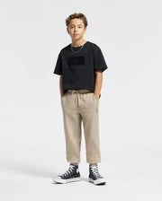 A young boy stands confidently with hands in his pockets, wearing a Psycho Bunny KIDS ROMEO HEAVY WEIGHT RELAXED FIT TEE in black, paired with beige pants and black high-top sneakers. The plain and minimal background highlights his casual yet stylish outfit.
