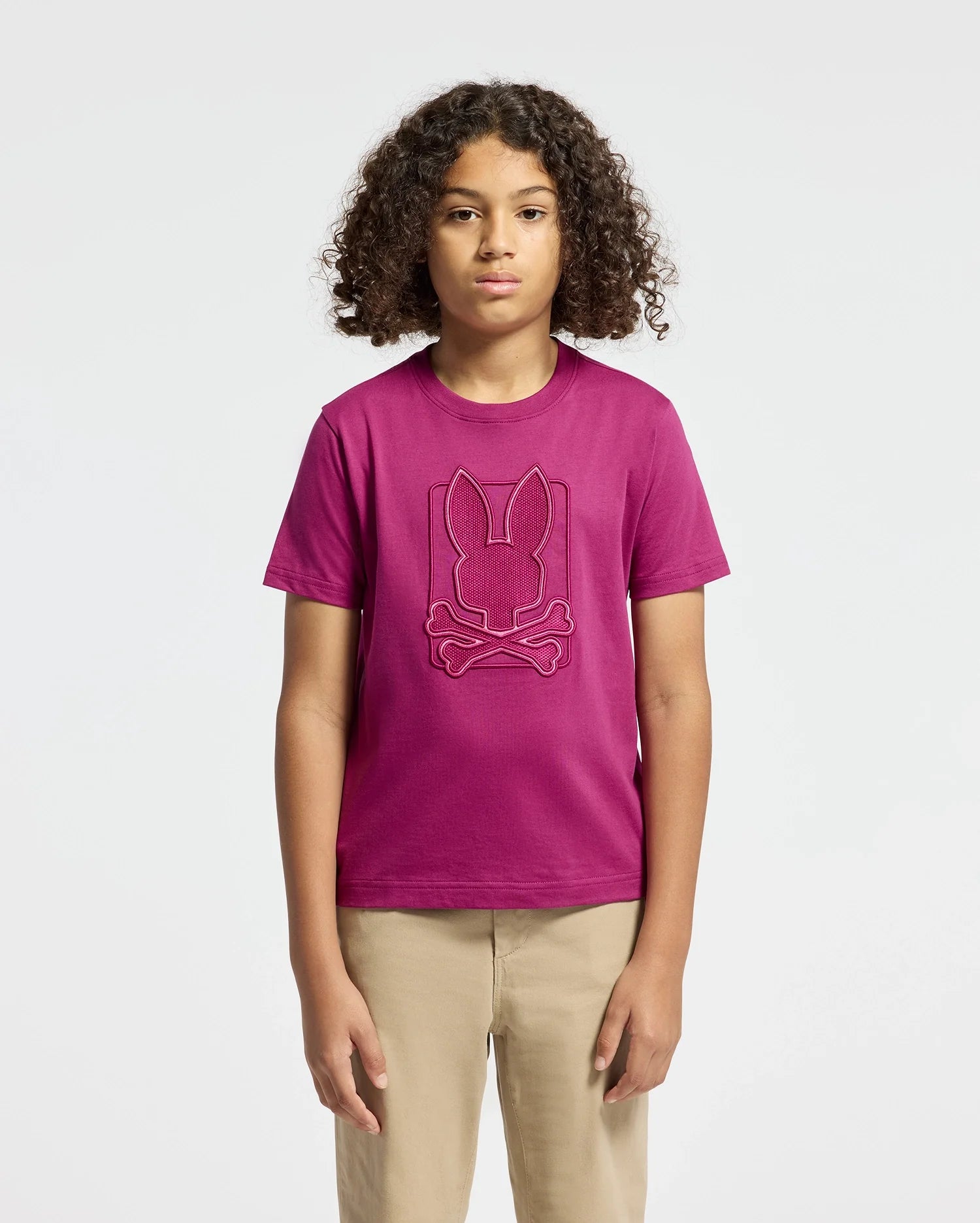 Against a simple background, a young person with curly hair is seen wearing the KIDS PIERCE GRAPHIC TEE - B0U115E200 by Psycho Bunny. This magenta T-shirt, crafted from Kids' Pima Cotton, features an embroidered bunny graphic logo above crossbones and is paired with beige pants.