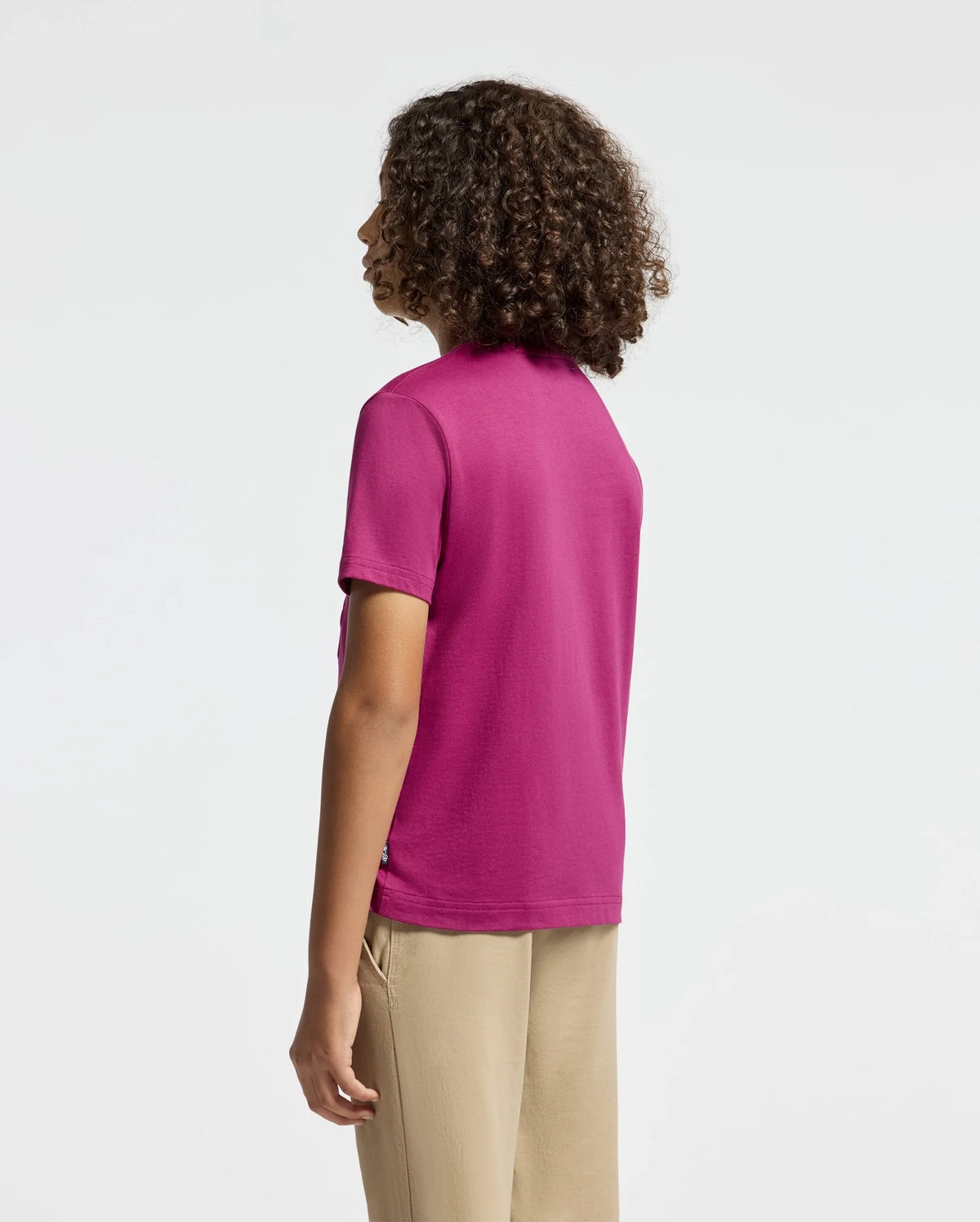 A person with curly hair is standing and facing away, wearing a bright pink KIDS PIERCE GRAPHIC TEE by Psycho Bunny featuring an embroidered bunny graphic, paired with beige pants against a plain, light background.