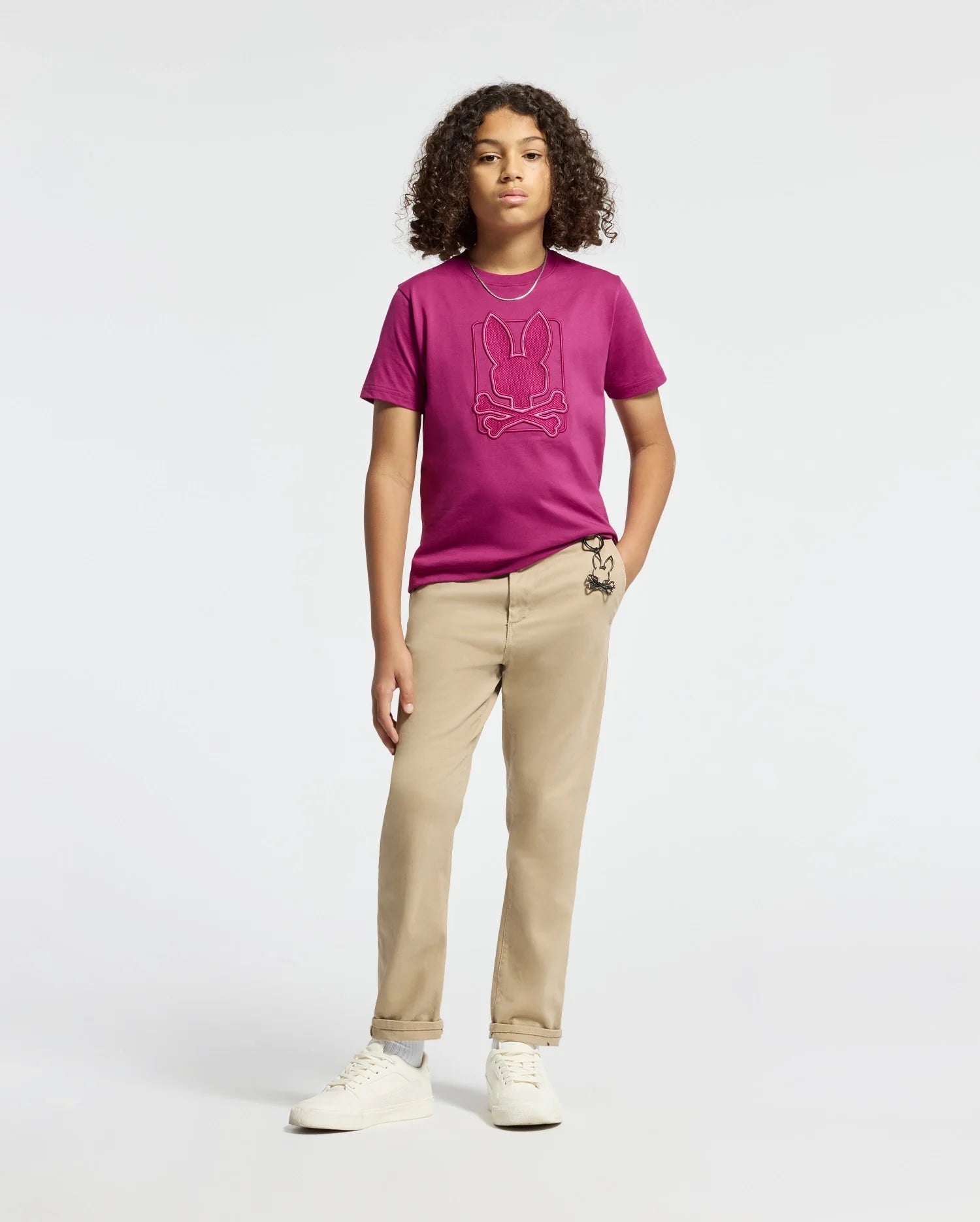 A young individual with curly hair exudes confidence in a KIDS PIERCE GRAPHIC TEE - B0U115E200 by Psycho Bunny, featuring a playful bunny design. They complement it with beige pants adorned with an embroidered small character design and finish the outfit with white sneakers. The backdrop is simple and light-colored.