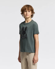 A young person with curly brown hair stands against a plain background, wearing the Psycho Bunny KIDS PIERCE GRAPHIC TEE - B0U115E200 in green and khaki pants. Made from kids' Pima cotton, the tee features a large bunny graphic that complements their neutral expression as they look at the camera.