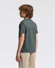 A young person with curly hair stands sideways, facing left. They wear a dark green, short-sleeved KIDS PIERCE GRAPHIC TEE by Psycho Bunny paired with light beige pants against a plain white backdrop.