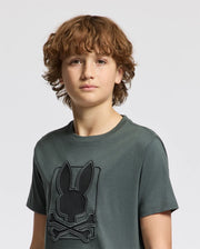A young person with curly hair is wearing a dark green KIDS PIERCE GRAPHIC TEE - B0U115E200 by Psycho Bunny, which features an embroidered design of a rabbit head and crossed bones. The background is a plain light gray.