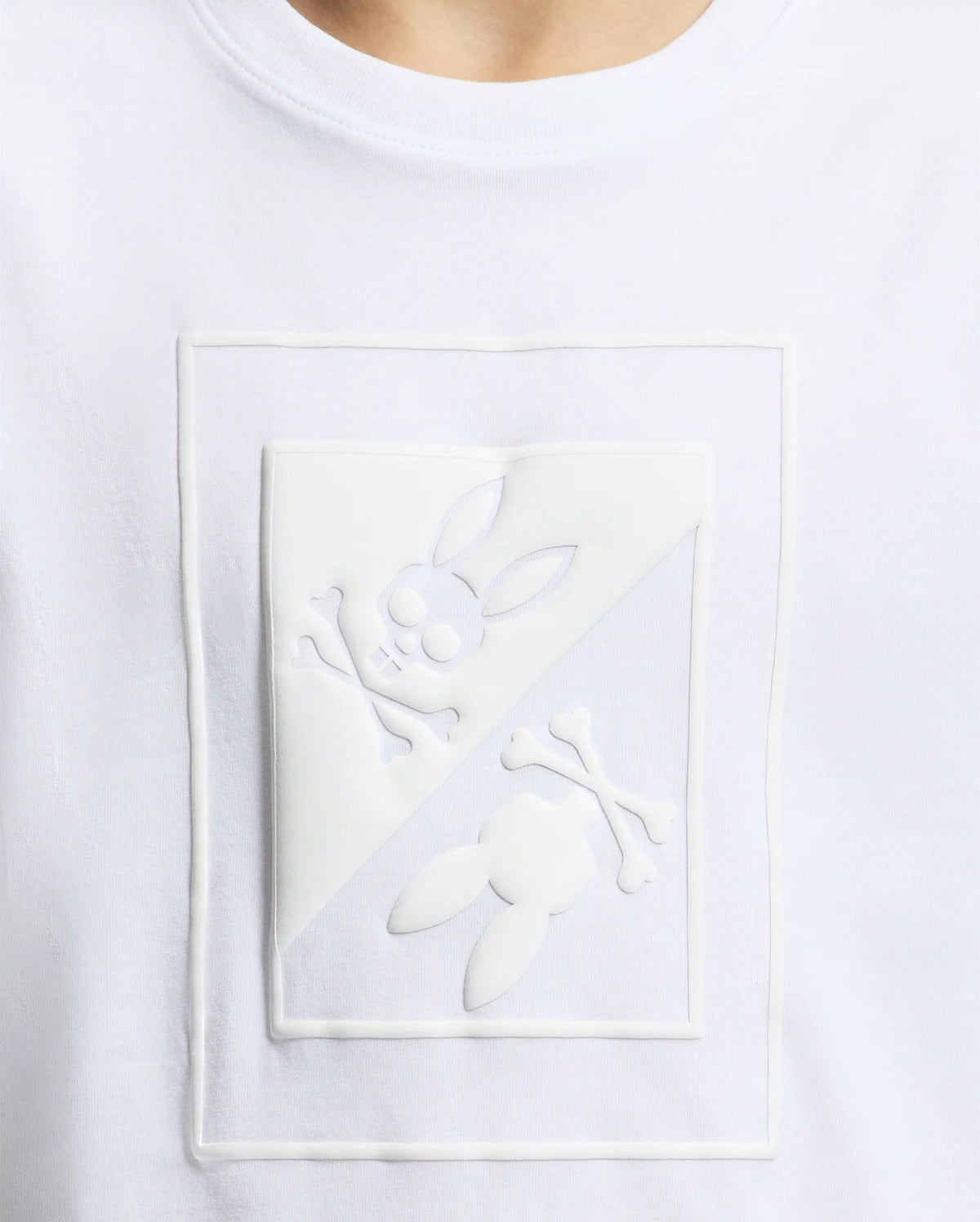 Close-up of the Psycho Bunny KIDS DOTSERO PRINTED GRAPHIC TEE - B0U197E200, a white children's shirt made from soft Peruvian Pima cotton. It features an eye-catching embossed puff print design that depicts a rabbit skull with crossbones and ears, split diagonally with one half upright and the other inverted.