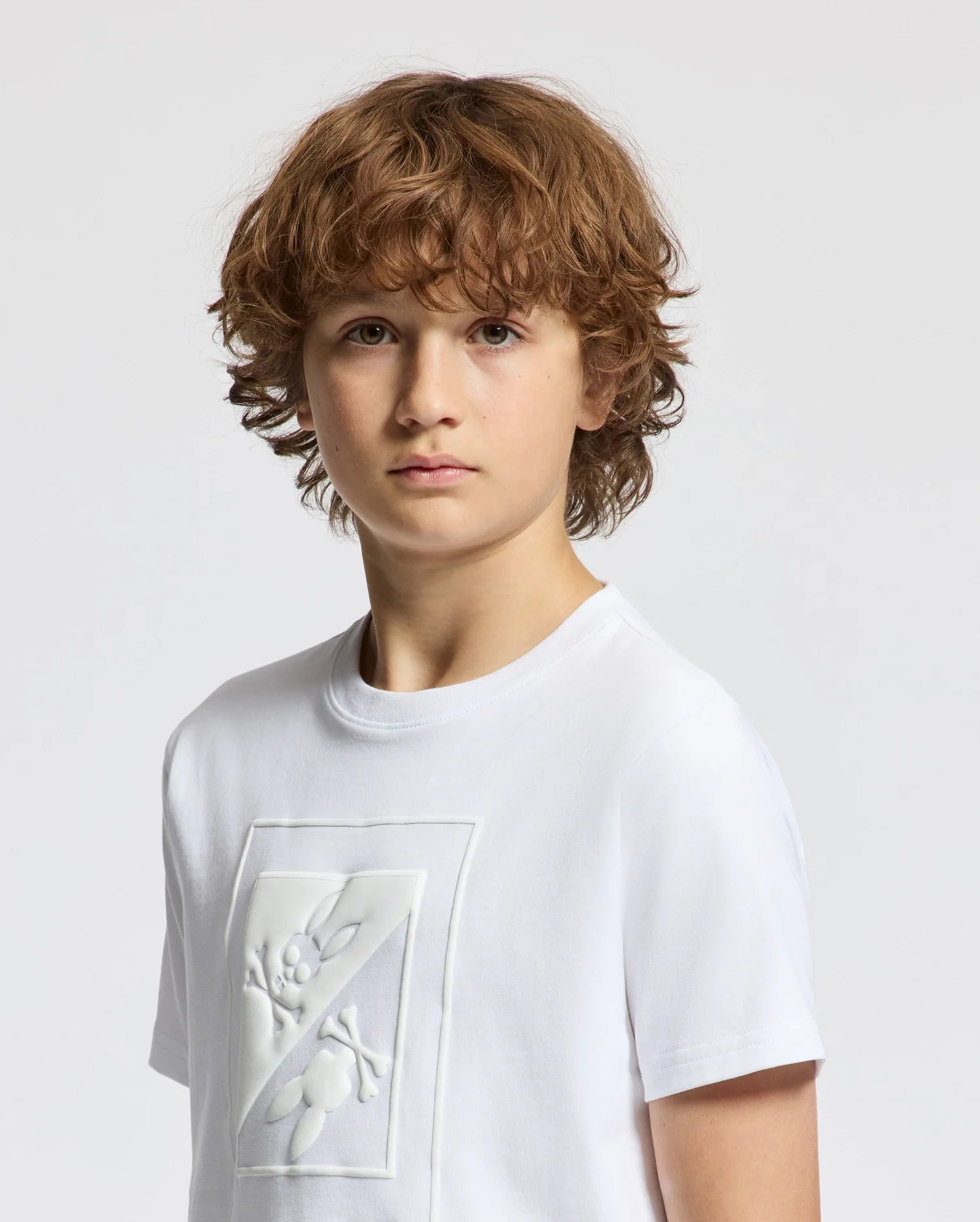 A young boy with curly brown hair wears the Psycho Bunny KIDS DOTSERO PRINTED GRAPHIC TEE in white, featuring a puff print graphic design on the front. Made from soft Peruvian Pima Cotton, he gazes at the camera with a neutral expression against a plain light background.