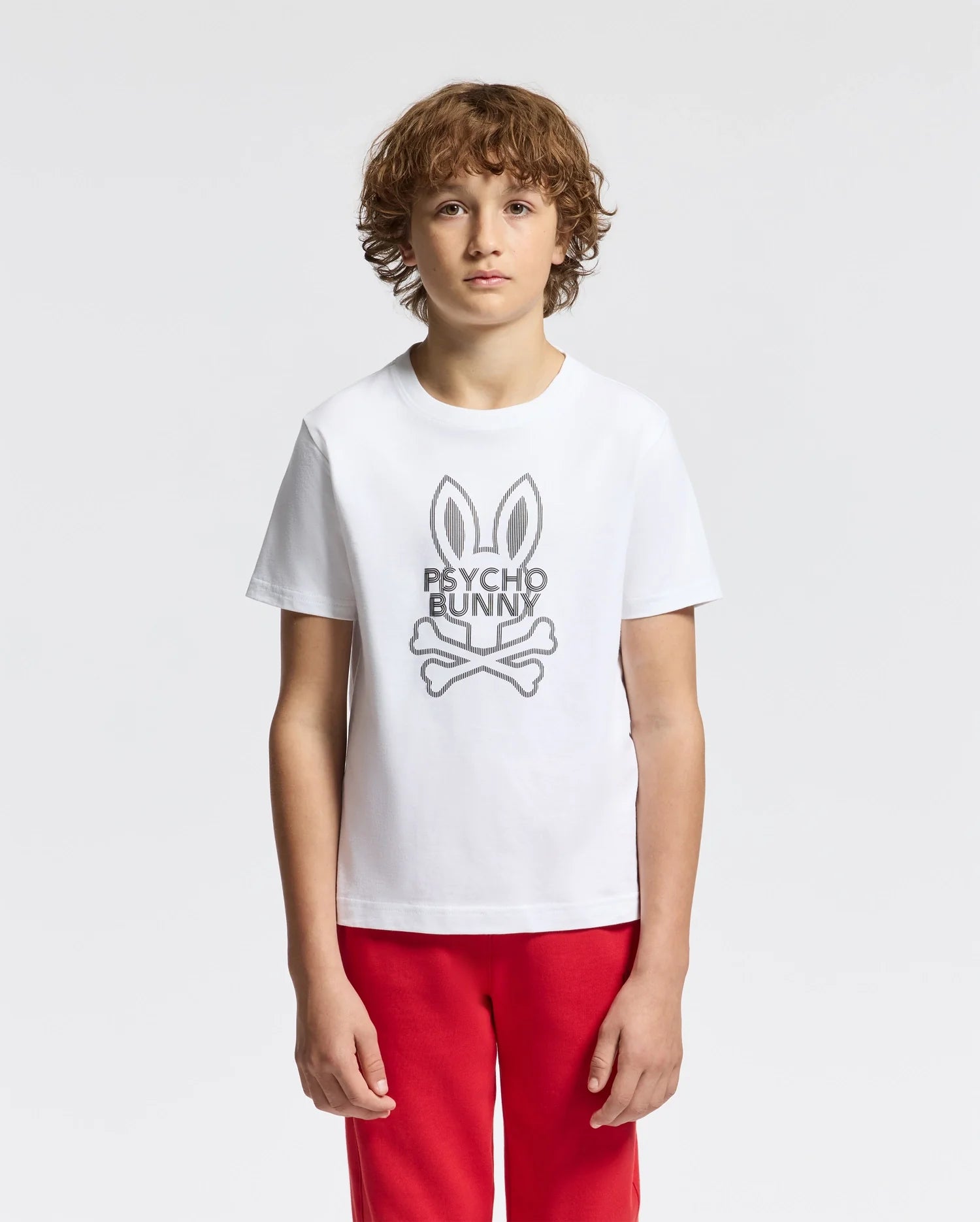 A young person with curly hair wears the KIDS DUMON GRAPHIC TEE - B0U271E200, made of Pima cotton, featuring a stylized bunny logo and "Psycho Bunny" text. They pair it with red pants against a light gray backdrop.