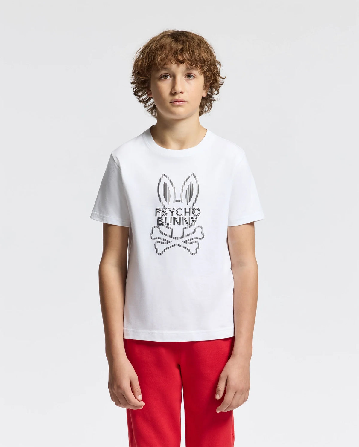 A young person with curly hair wears the KIDS DUMON GRAPHIC TEE - B0U271E200, made of Pima cotton, featuring a stylized bunny logo and "Psycho Bunny" text. They pair it with red pants against a light gray backdrop.