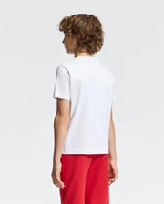 A person with curly brown hair is standing back, wearing a classic white Psycho Bunny KIDS DUMON GRAPHIC TEE - B0U271E200 and red pants against a light gray background.