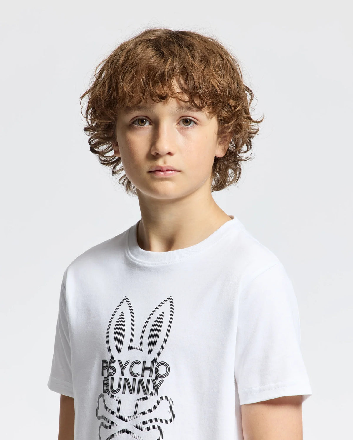A boy with curly brown hair wears a stylish Kids Dumon Graphic Tee (B0U271E200) made of soft Pima cotton, featuring a bunny and "Psycho Bunny" text. He stands against a plain white background, looking directly at the camera.