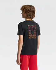 A person with curly hair is sporting the KIDS RIO BACK GRAPHIC TEE - B0U363E200, a trendy black graphic tee from Psycho Bunny, crafted from pima cotton and showcasing a vibrant bunny design along with the words "Psycho Bunny" on the back. This stylish look is complemented by red pants against a plain white background.