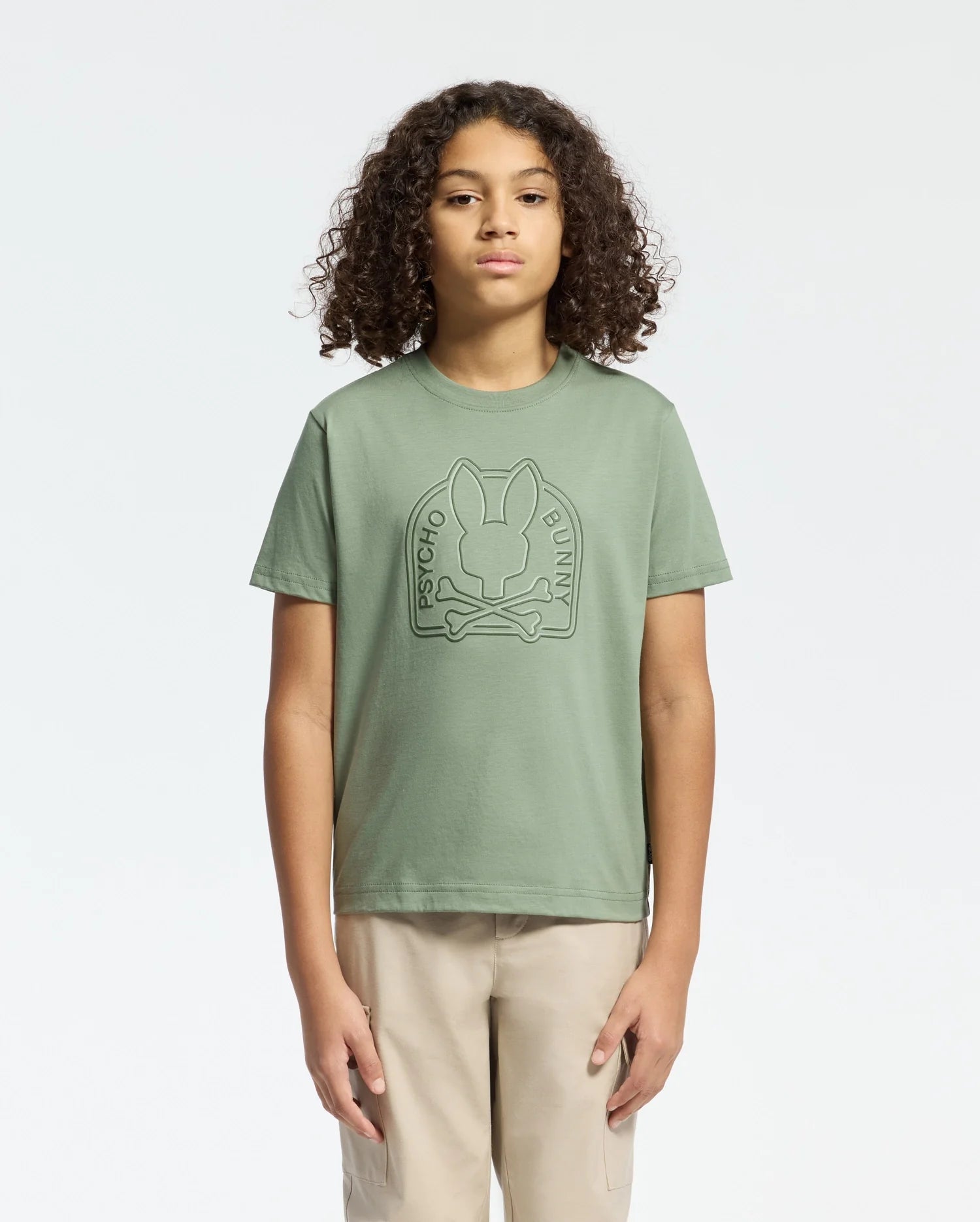 A person with curly hair wears a Psycho Bunny sage green KIDS MELBOURNE GRAPHIC TEE - B0U410F200, showcasing a large bunny and crossbones water-based print on the front. They pair it with beige pants against a plain white background.
