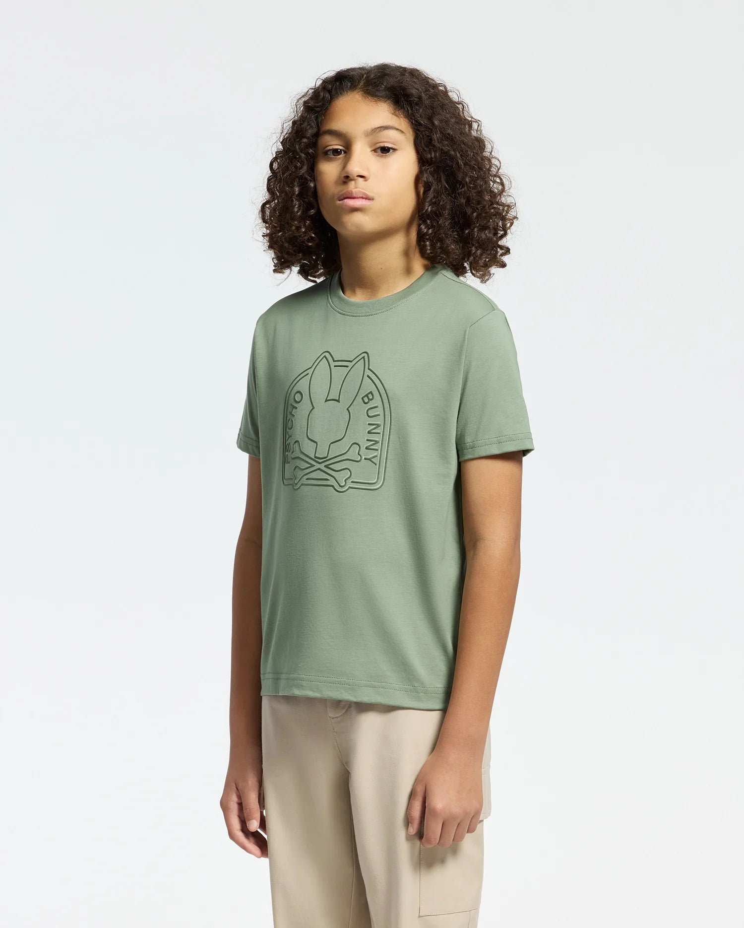 A curly-haired individual wears Psycho Bunny's KIDS MELBOURNE GRAPHIC TEE in sage green, made of soft Pima cotton with a bunny design. They pair it with beige pants against a white background.