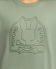 Close-up of the Psycho Bunny Kids Melbourne Graphic Tee in green Pima cotton, featuring a raised water-based print with a stylized bunny head atop crossed bones within an arch, with "Psycho Bunny" embossed on the arch's sides.