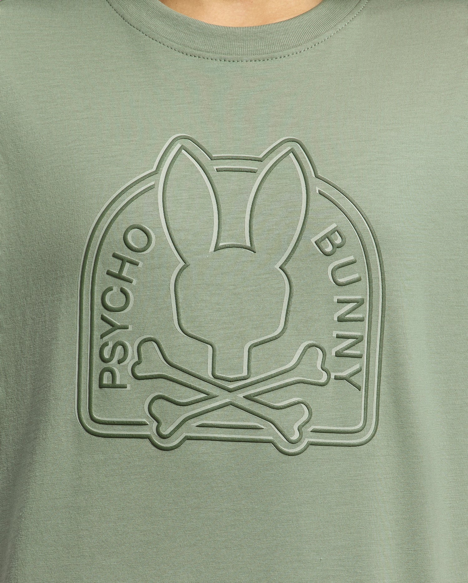 Close-up of the Psycho Bunny Kids Melbourne Graphic Tee in green Pima cotton, featuring a raised water-based print with a stylized bunny head atop crossed bones within an arch, with "Psycho Bunny" embossed on the arch's sides.