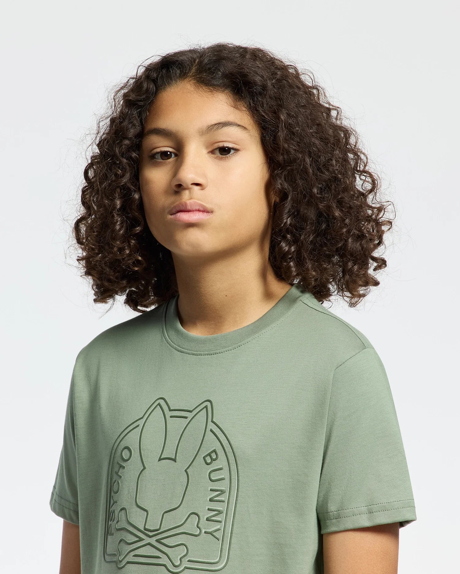 A young person with curly hair stands against a plain background wearing the Psycho Bunny KIDS MELBOURNE GRAPHIC TEE (B0U410F200), featuring an edgy, playful bunny graphic with crossed bones on a green Pima cotton t-shirt.