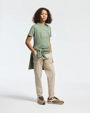 A person with curly hair wears Psycho Bunny's KIDS MELBOURNE GRAPHIC TEE in green with a water-based bunny print. They're styled in light beige cargo pants, brown sneakers, and have a matching green Pima cotton sweatshirt around their waist, against a plain light background.