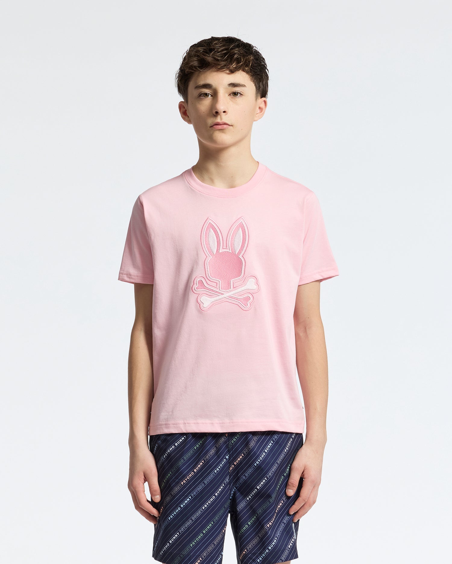A teen boy wears the Psycho Bunny KIDS NEW CASTLE EMBROIDERED GRAPHIC TEE (B0U415F200) in pink, made from soft Pima cotton with a bunny and crossbones design. He pairs it effortlessly with navy patterned shorts, standing against a plain white background.