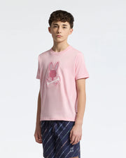 A young person with short, curly hair wears a pink Psycho Bunny KIDS NEW CASTLE EMBROIDERED GRAPHIC TEE, crafted from soft Pima cotton, showcasing a bunny and crossbones design. They pair it with dark blue patterned pants against a plain white backdrop.