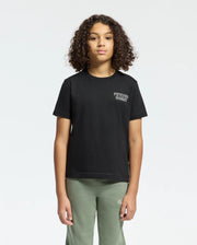A young person with curly hair stands against a plain background, wearing a Psycho Bunny black KIDS SYDNEY BACK GRAPHIC TEE in soft Pima cotton, featuring a small water-based logo on the chest and olive green pants. They have a neutral expression and maintain a regular fit, facing forward.