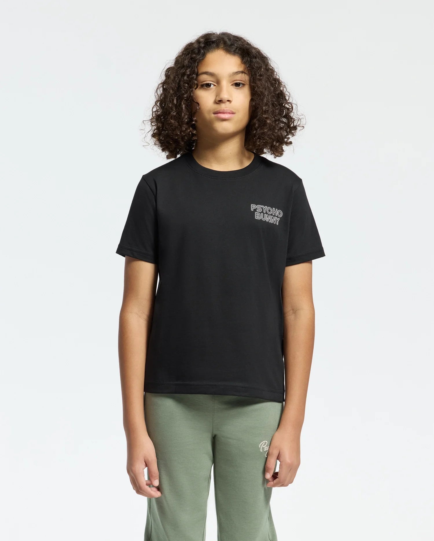 A young person with curly hair stands against a plain background, wearing a Psycho Bunny black KIDS SYDNEY BACK GRAPHIC TEE in soft Pima cotton, featuring a small water-based logo on the chest and olive green pants. They have a neutral expression and maintain a regular fit, facing forward.
