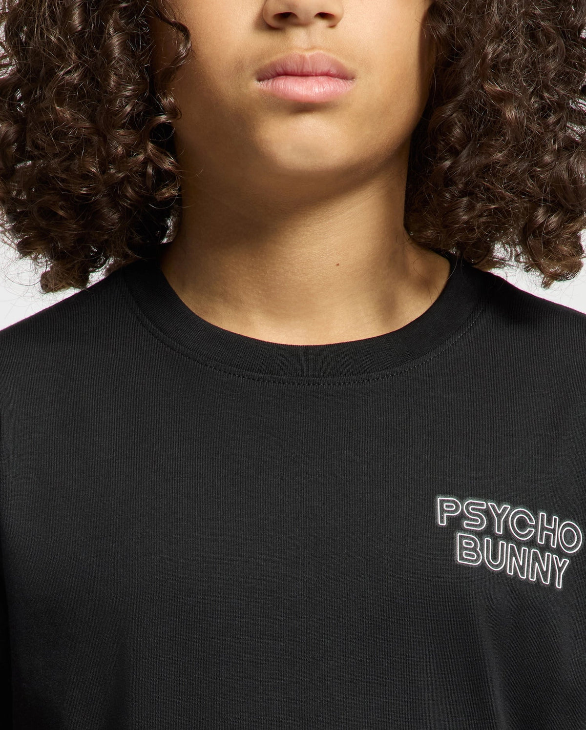 A person with curly hair wears the KIDS SYDNEY BACK GRAPHIC TEE, displaying "Psycho Bunny" in white on a black regular fit Pima cotton shirt. The subtle water-based print adds to its plain backdrop design.