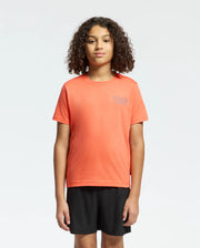 Against a plain background, a person with curly hair wears the KIDS SYDNEY BACK GRAPHIC TEE by Psycho Bunny in a regular fit, orange Pima cotton. The chest has small white water-based print text. They pair it with black shorts and gaze forward neutrally.