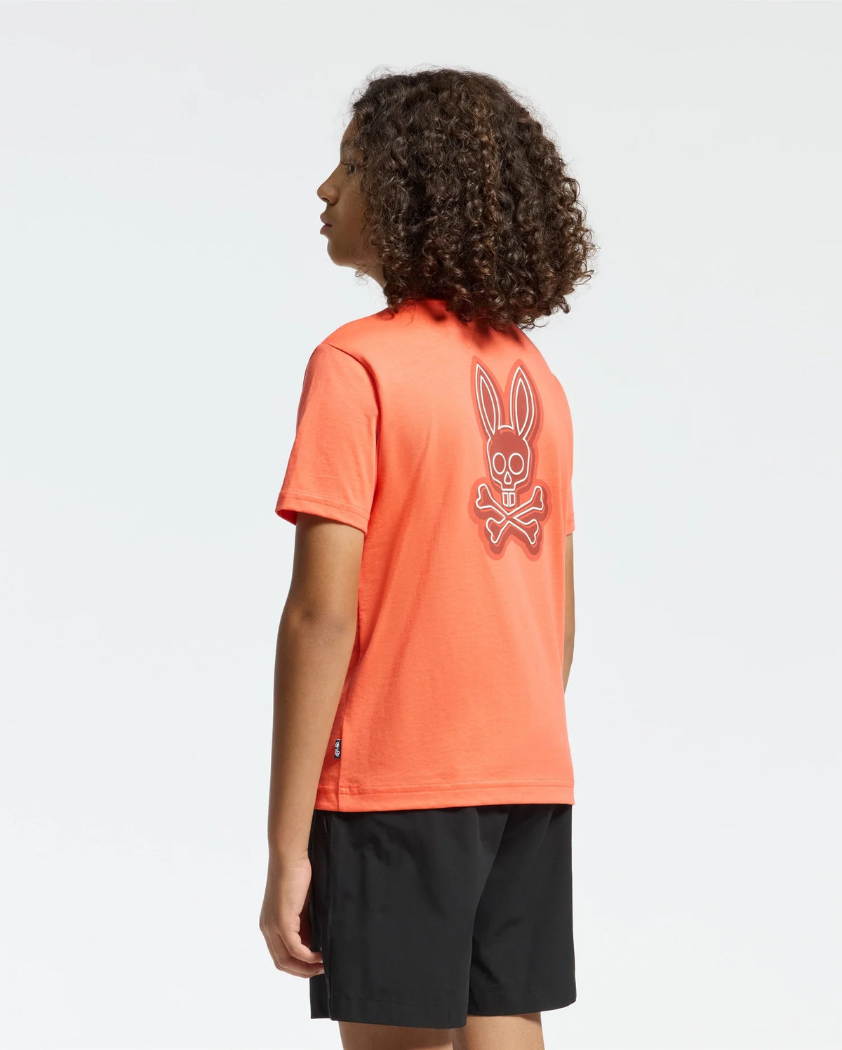 A person with curly hair stands sideways against a plain white background, wearing the Psycho Bunny KIDS SYDNEY BACK GRAPHIC TEE (B0U509F200) in regular fit orange Pima cotton, featuring a water-based bunny skull graphic on the back, paired with black shorts.