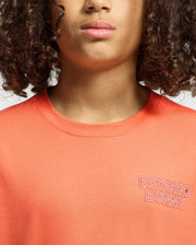 A person with curly hair wears the regular-fit orange "KIDS SYDNEY BACK GRAPHIC TEE" made of Pima cotton. It features a "Psycho Bunny" water-based print on the chest. The image focuses on the shirt and part of their face, emphasizing their closed lips.