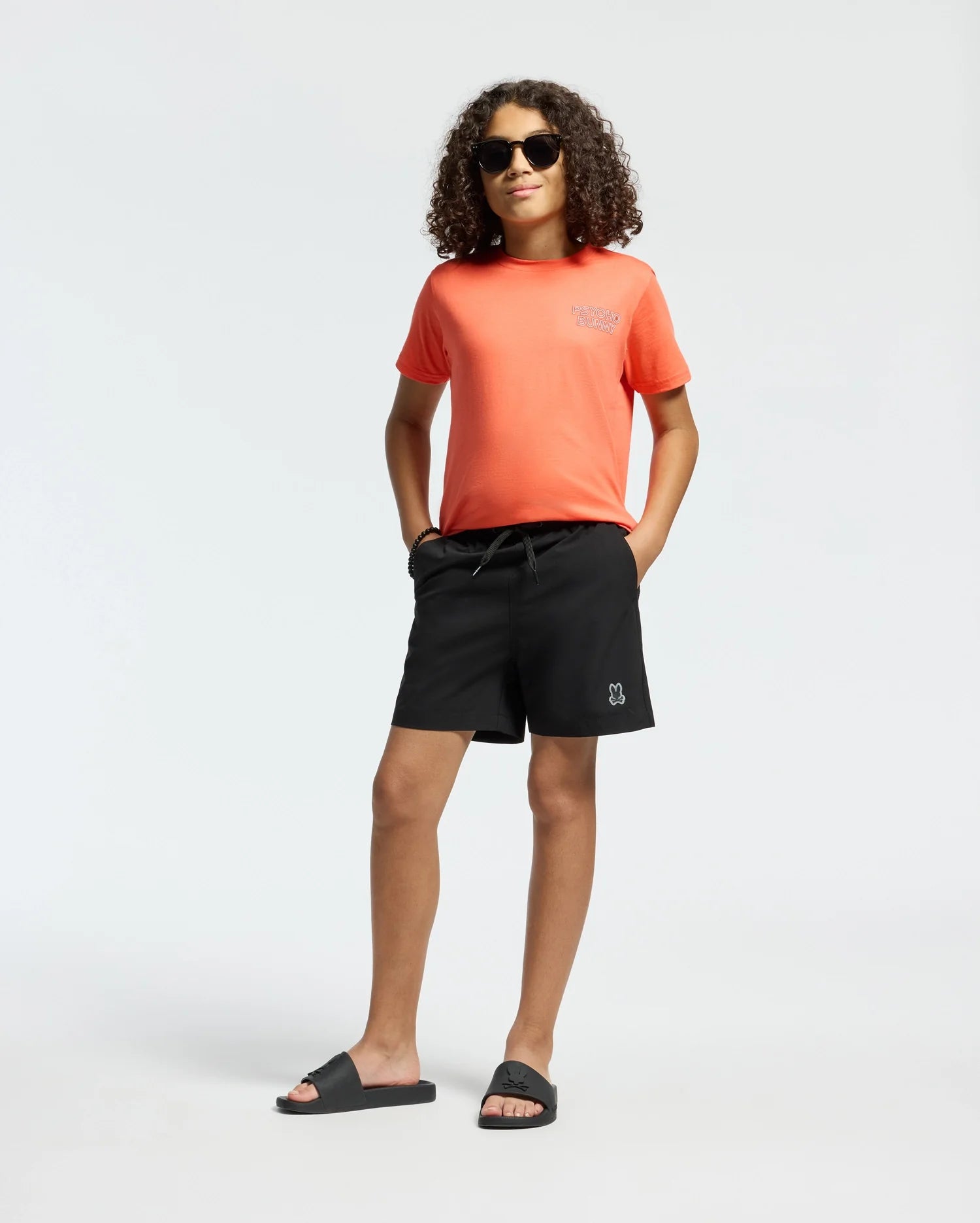 A person with curly hair, wearing sunglasses and a bright orange Psycho Bunny KIDS SYDNEY BACK GRAPHIC TEE in regular fit Pima cotton, black shorts, and black slide sandals, stands confidently with hands in pockets against a plain background.