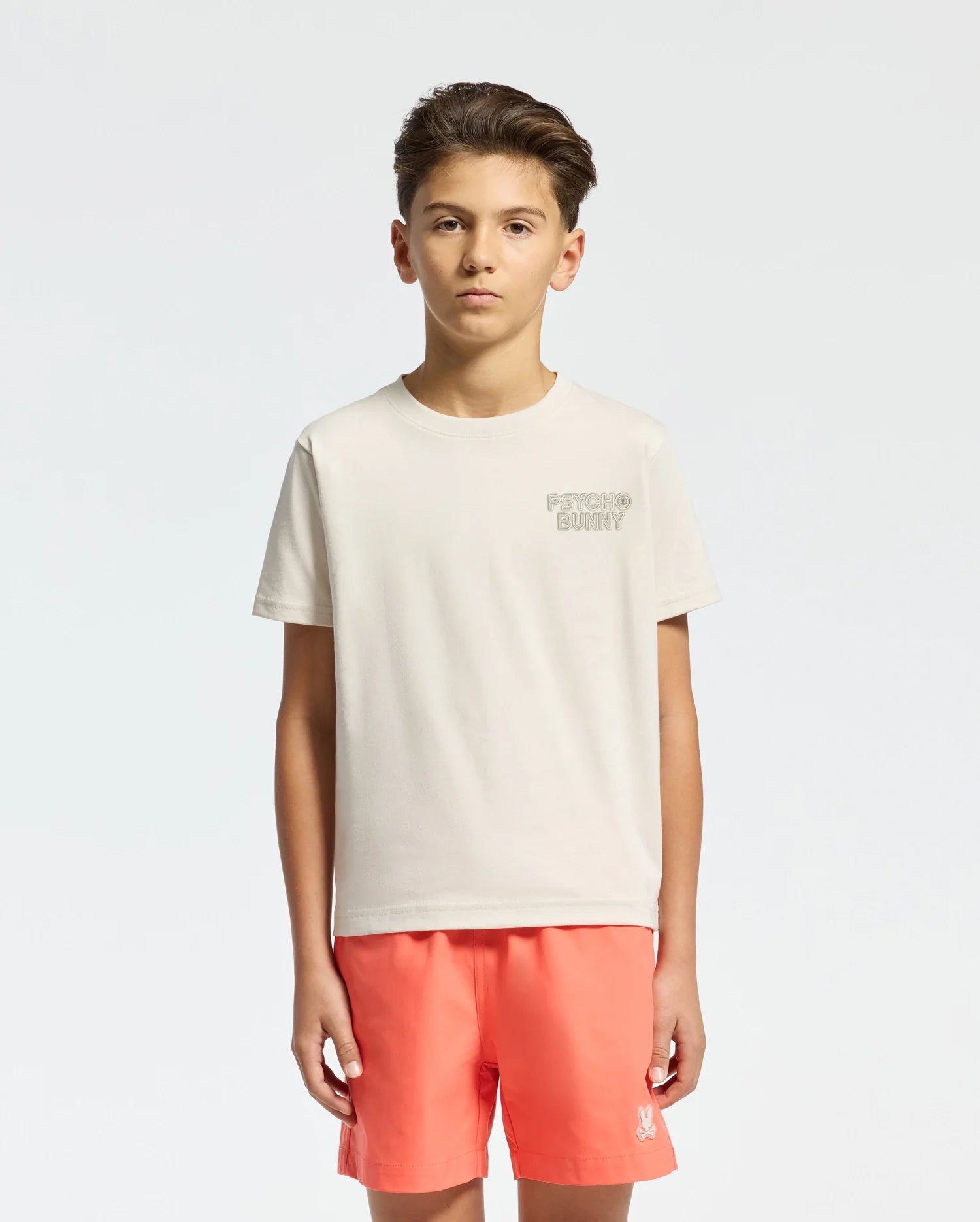 A young person with short brown hair stands against a white background, wearing a light beige KIDS SYDNEY BACK GRAPHIC TEE by Psycho Bunny, featuring subtle water-based print text on the chest, and bright coral shorts with a small logo.