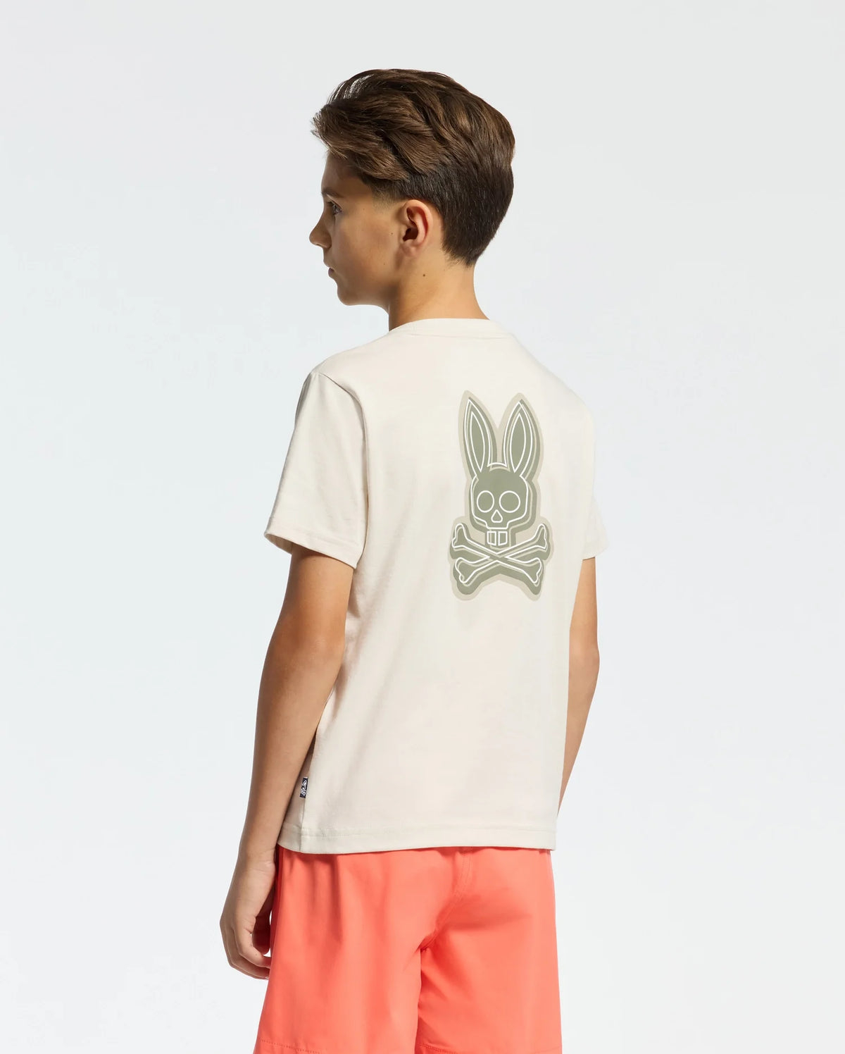 A boy with short hair stands sideways, wearing a Psycho Bunny KIDS SYDNEY BACK GRAPHIC TEE (B0U509F200) in beige Pima cotton with a bunny and crossed bones water-based print on the back, paired with bright coral shorts against a plain white background.