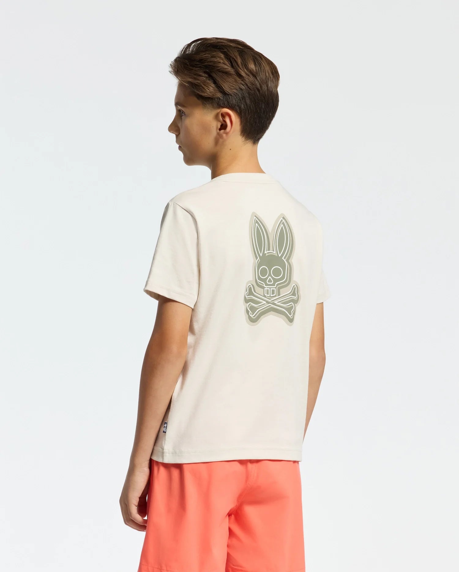 A boy with short hair stands sideways, wearing a Psycho Bunny KIDS SYDNEY BACK GRAPHIC TEE (B0U509F200) in beige Pima cotton with a bunny and crossed bones water-based print on the back, paired with bright coral shorts against a plain white background.