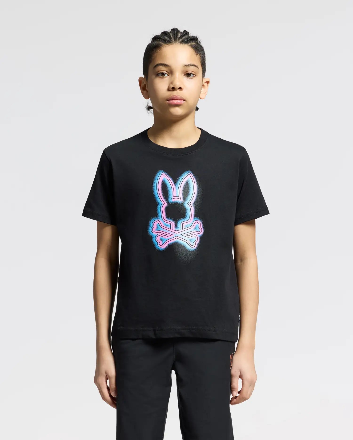 A youngster wears a Psycho Bunny KIDS ANDROS GRAPHIC TEE - B0U525F200, showcasing a neon-style bunny above crossbones. Made from 100% Pima Cotton in Peru, they stand against a light gray backdrop, gazing at the camera.