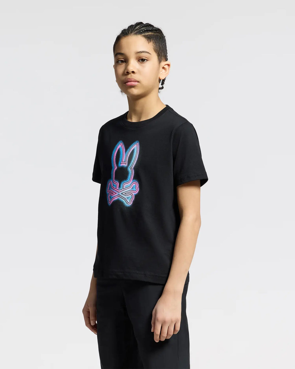A person with braided hair stands against a gray background, wearing the Psycho Bunny KIDS ANDROS GRAPHIC TEE (B0U525F200), featuring a neon-style bunny head and bones design. Made from 100% Pima Cotton in Peru, they gaze neutrally to the side.
