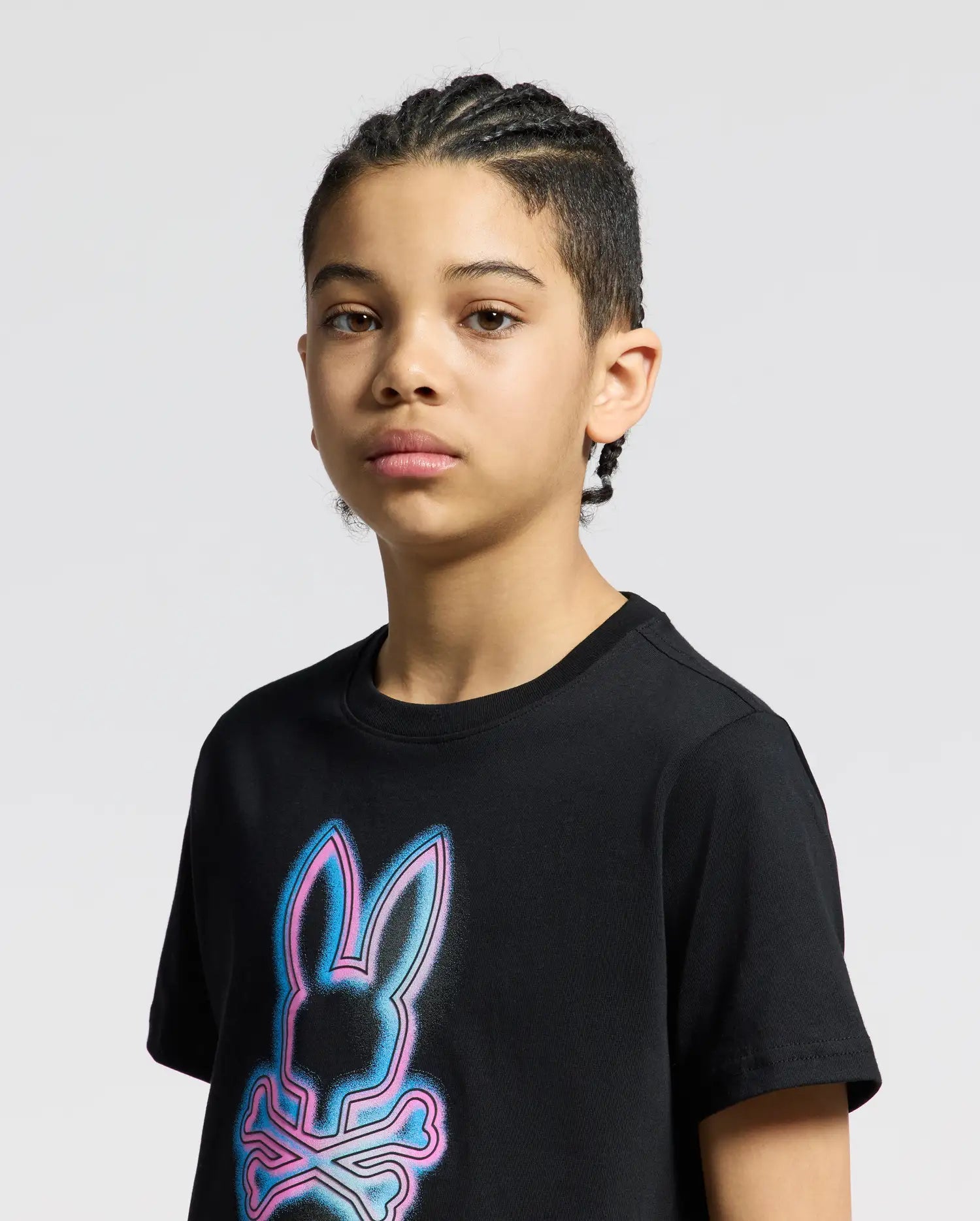A stylish young person with braided hair wears the Psycho Bunny KIDS ANDROS GRAPHIC TEE - B0U525F200, featuring a glowing bunny and crossbones design in black Pima Cotton. They maintain a neutral expression against a plain background.
